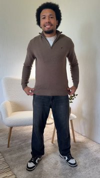 Brauner, langärmeliger Pullover mit Reißverschluss am Kragen und geripptem Saum, kombiniert mit dunklen Jeans und schwarz-weißen Turnschuhen. Strukturierter Hintergrund.