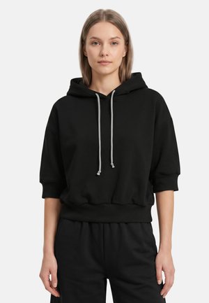 Vrouw met recht lichtbruin haar draagt een losse zwarte hoodie met korte mouwen en grijze trekkoorden, staand tegen een witte achtergrond.