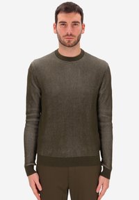 Uomo con capelli corti scuri e barba, che indossa un maglione a maniche lunghe a coste di colore verde oliva e pantaloni abbinati, in piedi con le braccia rilassate.