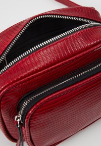 Borsa a tracolla rossa con texture, dotata di più scomparti, cerniere argento e tracolla regolabile. L'interno presenta un rivestimento nero.