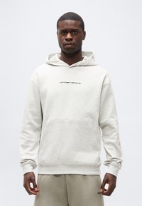 Νέος άνδρας μαύρου χρώματος δέρματος που φοράει ανοιχτό γκρι φούτερ Jordan Sport και μπεζ παντελόνι, στέκεται μπροστά σε λευκό, απλό φόντο.