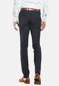 Marineblaue Hose aus glattem Stoff, slim fit, mit zwei Gesäßtaschen und einem dezenten Bundfalten vorne. Getragen mit einem braunen Gürtel und Schuhen.