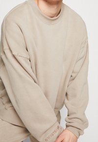 Felpa beige oversize con scollatura rotonda, polsini a coste e dettagli di cucitura in vista. Presenta un testo ricamato vicino all'orlo.
