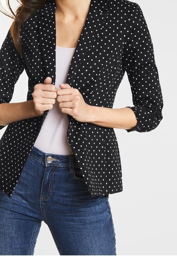 Femme portant un blazer noir à pois blancs par-dessus un haut blanc et un jean bleu, tenant le blazer fermé avec les deux mains.