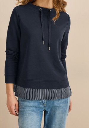 Woman wearing navy blue long-sleeve sweatshirt with drawstrings over a black-and-white striped shirt, paired with light blue jeans.