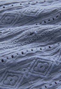 Textúrovaná tkanina v jemnej modrej farbe s prešívanými diamantovými vzormi a malými výrezmi. Materiál sa javí ako pletený s jemným, vlnitým dizajnom.