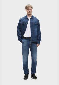 Τζάκετ τζιν με μπροστινές τσέπες, μπλε τζιν και μαύρα αθλητικά παπούτσια. Το τζάκετ έχει κλασική γραμμή, ενώ τα τζιν είναι στενά με ξεθωριασμένο φινίρισμα.