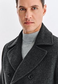 Szary wełniany płaszcz w wzór jodełki z szerokimi klapami, podwójnymi guzikami i teksturowanym wykończeniem, noszony na jasnoszarym swetrze z golfem.