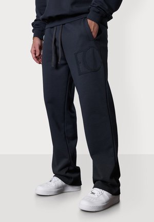 Navy Sweatpants mit einem entspannenden Schnitt, ausgestattet mit einem großen gestickten Logo auf dem linken Oberschenkel und einem verstellbaren Kordelzugbund; kombiniert mit weißen Sneakers.