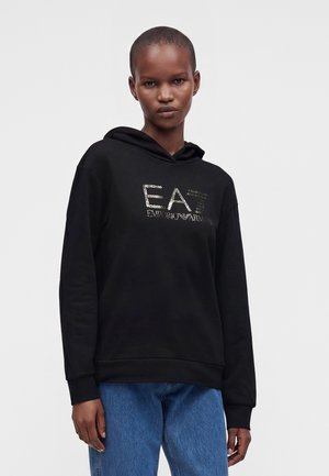 Person mit kurzen Haaren, die einen schwarzen Hoodie mit dem EA7 Emporio Armani Logo und blaue Jeans vor einem einfarbigen hellen Hintergrund trägt.