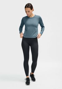 Lichte, lange mouwen, teal sporttop gecombineerd met zwarte, aansluitende leggings en zwarte sportschoenen, staand op een eenvoudige achtergrond.