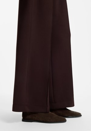 Braune Hosen mit weitem Bein, die dunkelbraune Wildleder flache Schuhe mit abgerundeten Zehen bedecken.