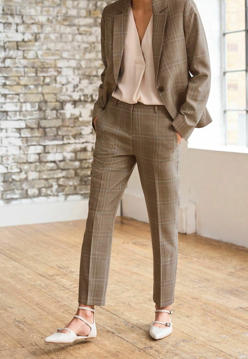 Femme portant un tailleur à carreaux beige avec un pantalon fuselé et une veste assortie, associé à un chemisier beige clair et des ballerines beige à brides à la cheville.
