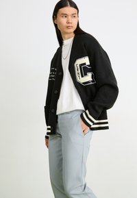 Cardigan tricotat negru cu dungi albe, nasturi frontali și aplicații. Purtați peste un top alb și asortat cu pantaloni gri deschis.