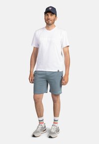 Biała bawełniana koszulka z logo "ROSSIGNOL", jasnoniebieskie szorty, skarpetki w wielokolorowe paski i szare buty sportowe. Ciemna czapka z logo.