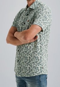 Camisa de manga corta hecha de tela ligera con un patrón de hojas en verde y blanco, cuenta con cuello, botones y un pequeño detalle de logo en el pecho.