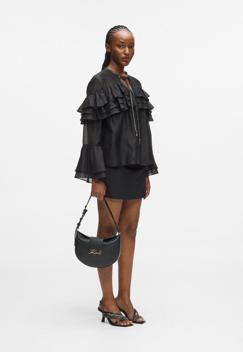 Γυναίκα με μαύρη ριγέ μπλούζα και φούστα, κρατώντας μια μαύρη τσάντα με χρυσό λογότυπο, και φορητή μαύρες γόβες με λεπτομέρεια φράντζας.