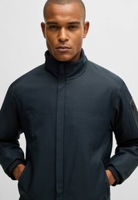 Dunkelblaue Jacke mit hohem Kragen, strukturiertem Stoff, zwei Brusttaschen und einem Akzentpatch am linken Arm.