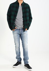 Zeleno-černá kostkovaná flanelová košile, šedé tričko s výstřihem do V pod tím, světle modré džíny a černé tenisky s bílou podešví.