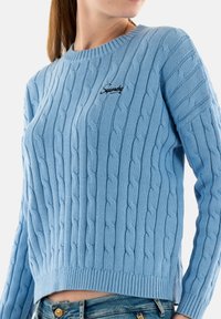 Suéter de punto trenzado azul claro con cuello redondo, dobladillo acanalado y un logo bordado en la parte superior izquierda del pecho.