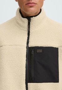 Beige Fleecejacke mit hohem Kragen, schwarzer Brusttasche mit Reißverschluss und Logo-Patch. Weicher Stoff mit kontrastierenden Farbdetails.