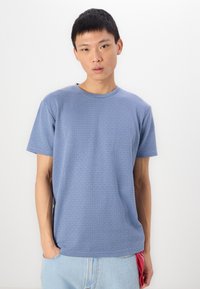 Μπλε κοντομάνικο μπλουζάκι με υφή πλέγματος, στρογγυλή λαιμόκοψη, χαλαρή εφαρμογή, σε συνδυασμό με ανοιχτόχρωμες τζιν σορτς.