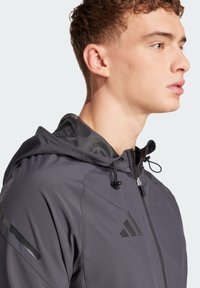Grijze hoodie met een strakke, gladde textuur. Voorzien van verstelbare koordjes en zwarte accenten, waaronder een embleem op de borst.