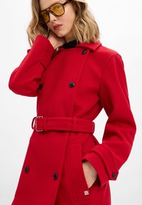 Cappotto rosso in lana con design doppiopetto, bottoni neri, vita con cintura e tasche laterali con dettaglio del logo. Tessuto texturizzato e colletto.