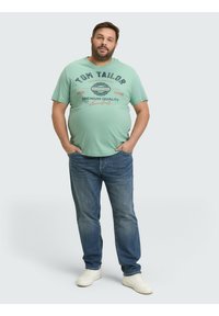 Hellgrünes T-Shirt mit schwarzem und rotem Text, aus Baumwolle, runder Ausschnitt, kombiniert mit blauen Jeans und weißen Turnschuhen, einfarbiger Hintergrund.