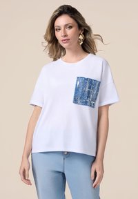 Camiseta blanca de manga corta con un bolsillo texturizado azul, con un patrón azul y plateado. Corte relajado, combinada con jeans de color azul claro.