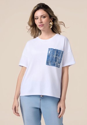 Witte t-shirt met korte mouwen, voorzien van een gestructureerde blauwe zak met een blauw- en zilverpatroon. Ontspannen pasvorm, gecombineerd met lichtblauwe jeans.