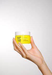 Balm detergente giallo in un barattolo bianco con coperchio, etichettato "NIP+FAB Ceramide Fix," tenuto da una mano su uno sfondo chiaro.