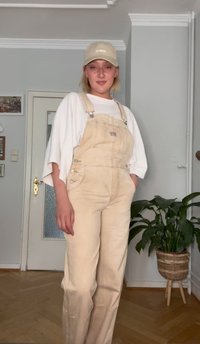 Beige overalls állítható pántokkal és oldalsó gombokkal, egy fehér, túlméretezett pólóval és egy világos bézs sapkával párosítva. Beltéri környezet növényekkel.