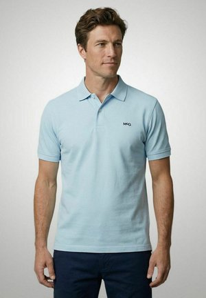 Man draagt een lichtblauw poloshirt met korte mouwen en kraag en donkerblauwe broek, staande tegen een effen lichte achtergrond.