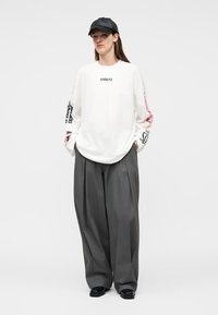 Frau trägt ein weißes Langarmshirt mit Aufschrift, weite graue Hose, schwarze Mütze und schwarze Schuhe, steht mit den Händen in den Taschen.