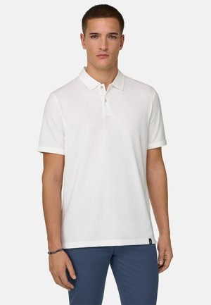 Jeune homme portant un polo blanc uni à manches courtes et un pantalon bleu, debout face à l'avant sur un fond blanc.