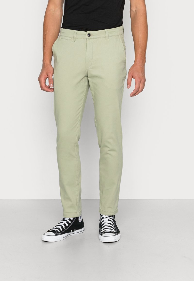 Homme portant un pantalon slim vert clair et des baskets montantes noires debout sur un fond blanc.