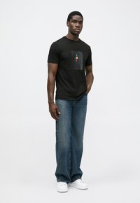 Homme portant un t-shirt graphique noir, un jean bleu ample et des baskets blanches, se tenant avec une main dans la poche devant un fond uni.