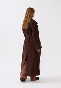 Brauner langer Mantel mit einem Gürtel, aus strukturiertem Stoff, mit einem weiten Kragen und einem seitlichen Schlitz. Über hellrosa Hosen und dunklen Schuhen getragen.