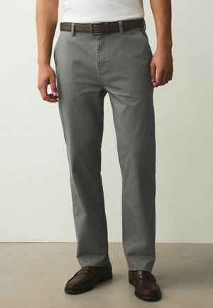 Man wearing gray straight-leg pants, brown leather belt, and dark brown loafers, standing against a plain light background.