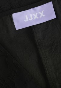 Juoda tekstūruota medžiaga su sulankstytu purpuriniu etiketu, ant kurio užrašyta "JJXX". Kraštai apdoroti juodais siūlų siūlais, išryškinant medžiagos detales.