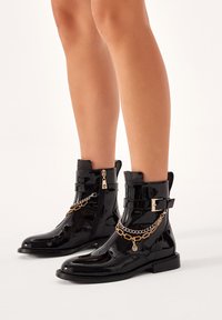 Botas de tobillo de charol negro con cierre de cremallera lateral, correas ajustables y un detalle de cadena dorada. Suela plana con punta redondeada.