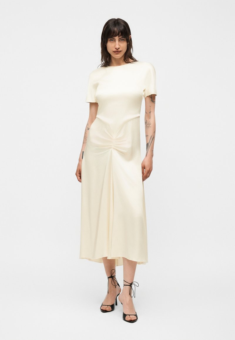 Femme debout portant une robe midi écrue à manches courtes avec un détail froncé à la taille, associée à des talons ouverts noirs avec liens à la cheville.