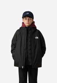 Černá venkovní bunda na zip s kapucí, nošená přes vínový péřový top. Na levé straně hrudi má bílý logo.