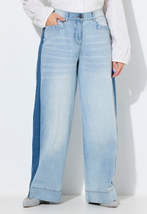 Kobieta ubrana w szerokie, jasnoniebieskie dżinsy z ciemniejszymi niebieskimi panelami po bokach, w połączeniu z białą bluzką z długim rękawem i szarymi butami.