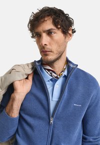 Blauer strukturierter Zip-Pullover über einem hellblauen Polohemd. Ein gemusterter Schal ist sichtbar; der Pullover hat ein Logo auf der Brust.