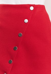 Falda envolvente roja hecha de una tela texturizada con un diseño diagonal. Presenta botones de presión de metal plateado a lo largo del lado frontal.