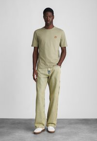Olive green cotton t-shirt with a small embroidered animal logo, paired with light yellow vertical-striped jeans and white sneakers.