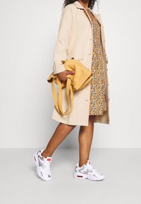 Beige trenchcoat over en blomsterkjole, bærer en sennepsgul taske. Hvide sneakers med grå accenter og røde detaljer fuldender looket.
