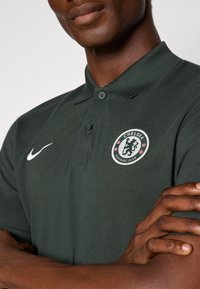 Mørkegrøn polo shirt med struktureret stof, der har et hvidt Nike-logo og et rundt Chelsea FC-skjold med røde og sorte detaljer.